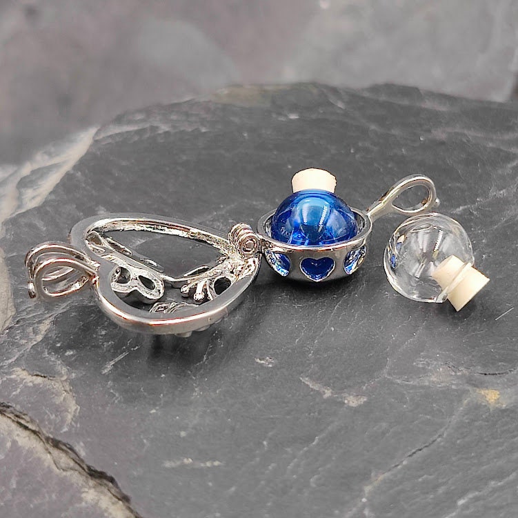 Geöffneter silberner Herz-Käfig mit Schmetterlingsmotiv auf Schieferstein, blaue befüllbare Glasperle mit Korkenstopfen und leere Ersatz-Glasperle daneben – Gedenkschmuck von Zinou Minimanufaktur