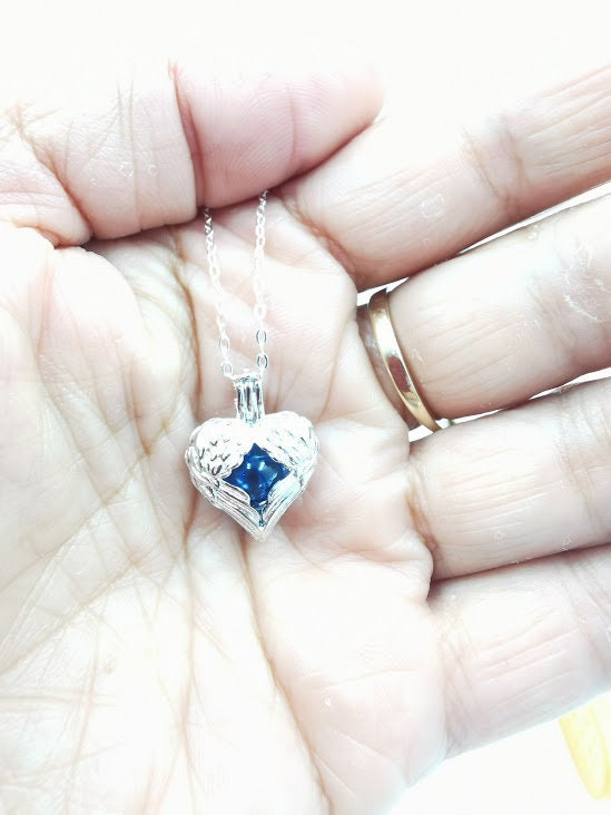 Silberner Herz-Käfig-Anhänger mit Engelflügeln und blauer Glasperle in der Handfläche gehalten, Nahaufnahme – DIY Gedenkschmuck von Zinou Minimanufaktur