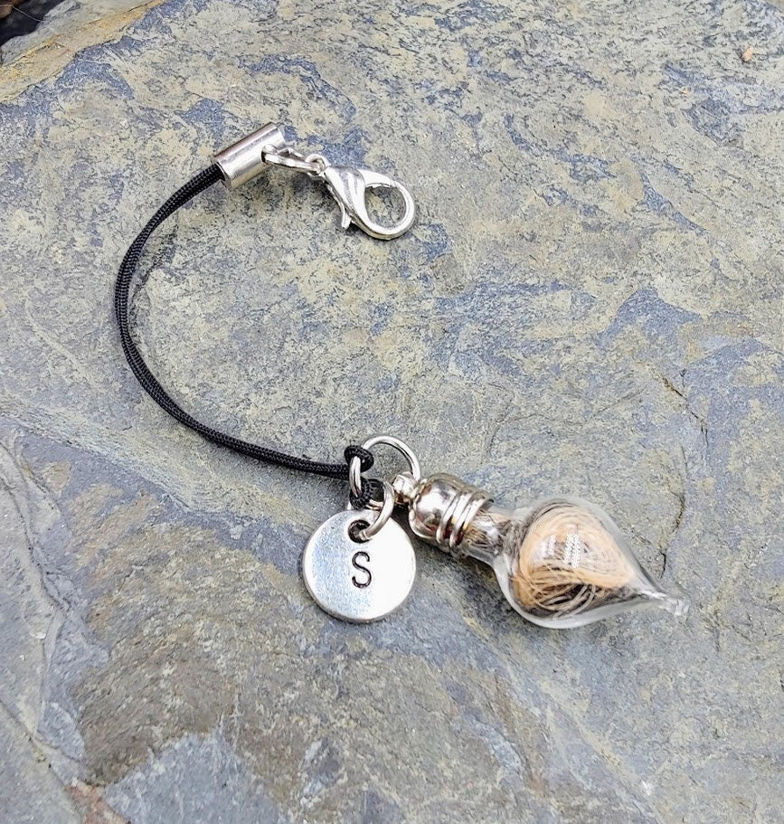 eine Glasphiole mit Tierhaar und silbernem Buchstaben-Charm S als Schlüsselanhänger an schwarzer Kordel auf Stein – personalisierter Gedenkschmuck von Zinou
