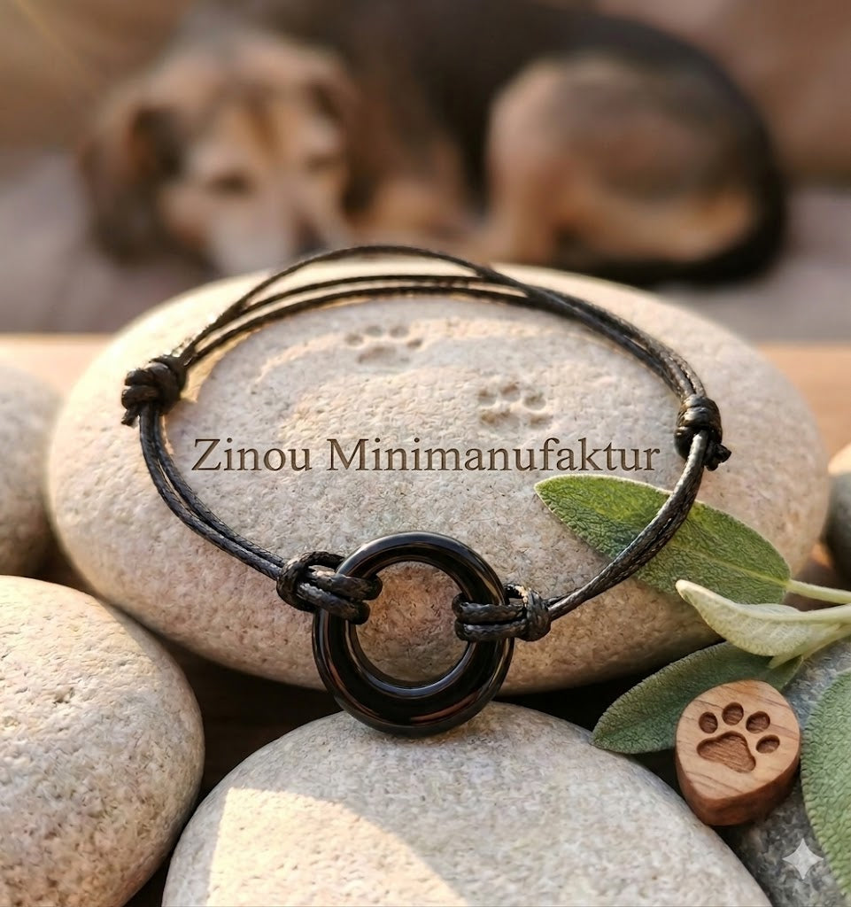 Angelo-Aschearmband mit schwarzem Ring-Element an schwarzer Kordel auf Kieselsteinen, Hund im Hintergrund – Gedenkschmuck von Zinou Minimanufaktur