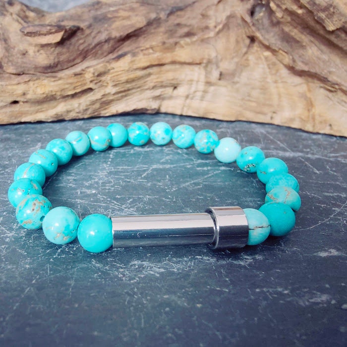 Türkis-Perlenarmband mit silbernem Miniurnen-Zylinder auf Schieferstein, Treibholz im Hintergrund – Gedenkschmuck DIY von Zinou Minimanufaktur