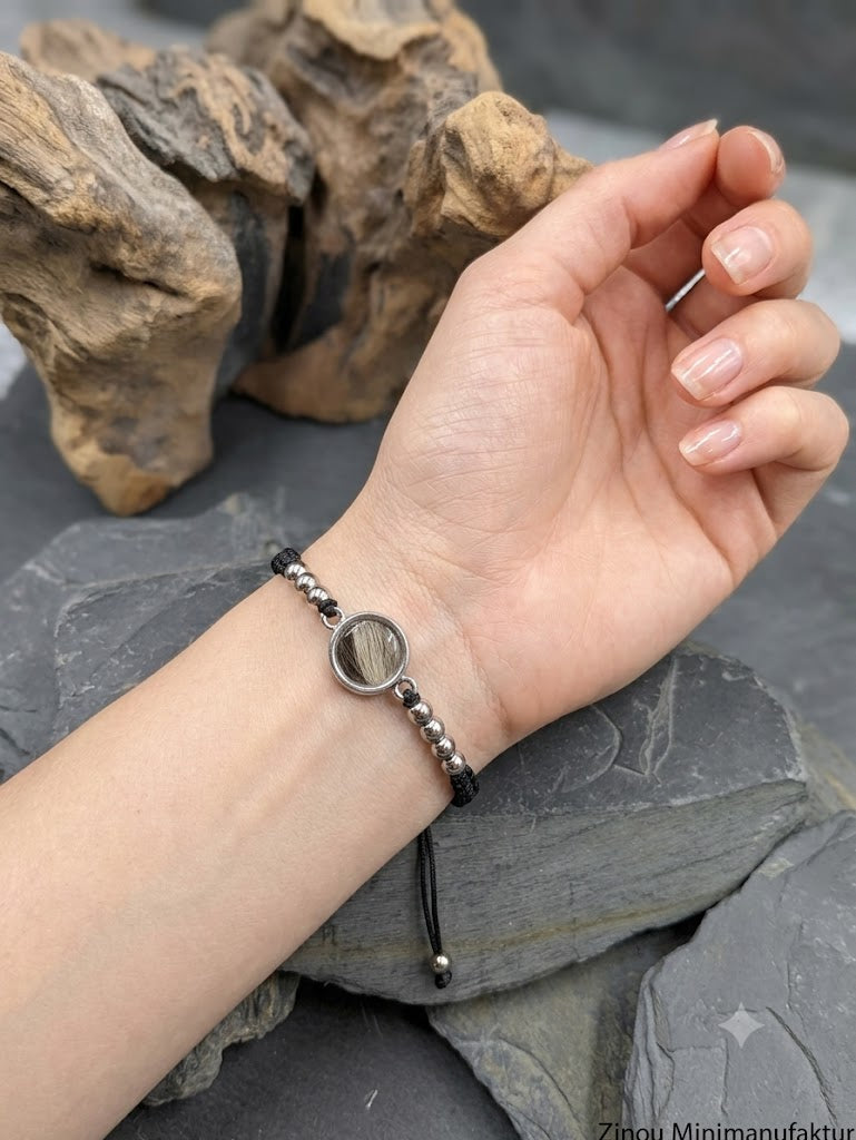 Schwarzes Makramee-Armband mit einer runden Fassung aus eingearbeitetem Tierhaar und silbernen Edelstahlperlen, getragen am Handgelenk.