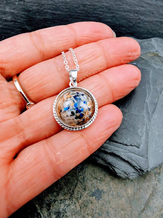 Silberner Dahlia-Anhänger mit weißem Asche-Einschluss und blauen Partikeln in einer Handfläche gehalten auf Schieferstein – Ascheschmuck von Zinou Minimanufaktur