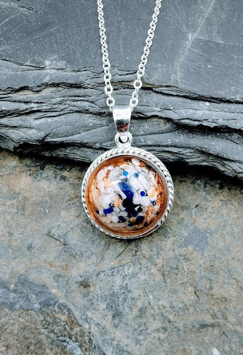 Silberner Dahlia-Anhänger mit weißem Asche-Einschluss, blauen Partikeln und Goldflitter an Silberkette auf Schieferstein – Ascheschmuck von Zinou Minimanufaktur