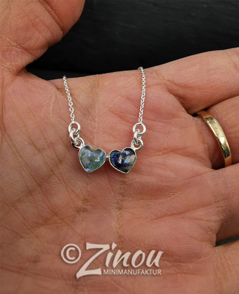 Zwei silberne Herzanhänger mit blau-grünem Asche-Einschluss in der Hand gehalten – Gedenkschmuck von Zinou Minimanufaktu