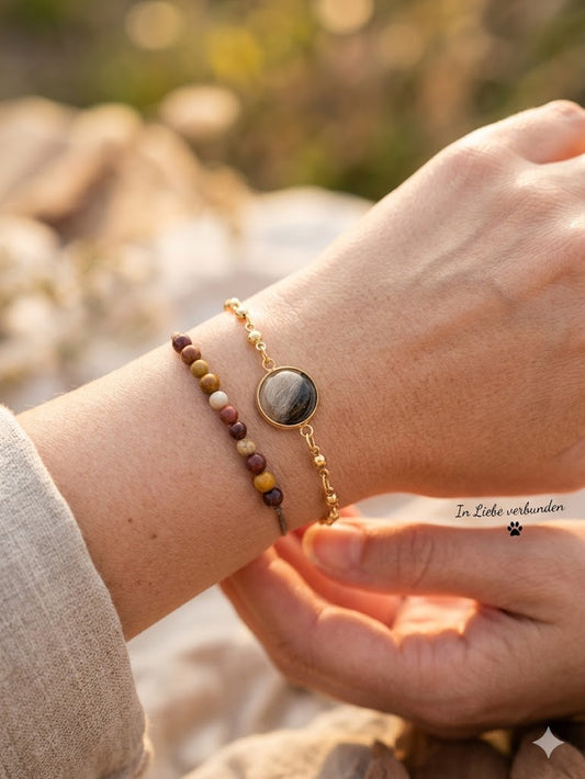 Hand trägt ein feines filigranes Goldarmband mit einer runden Perle gefüllt mit Haaren vom eigenen Haustier, kombiniert mit einem bunten Perlenarmband