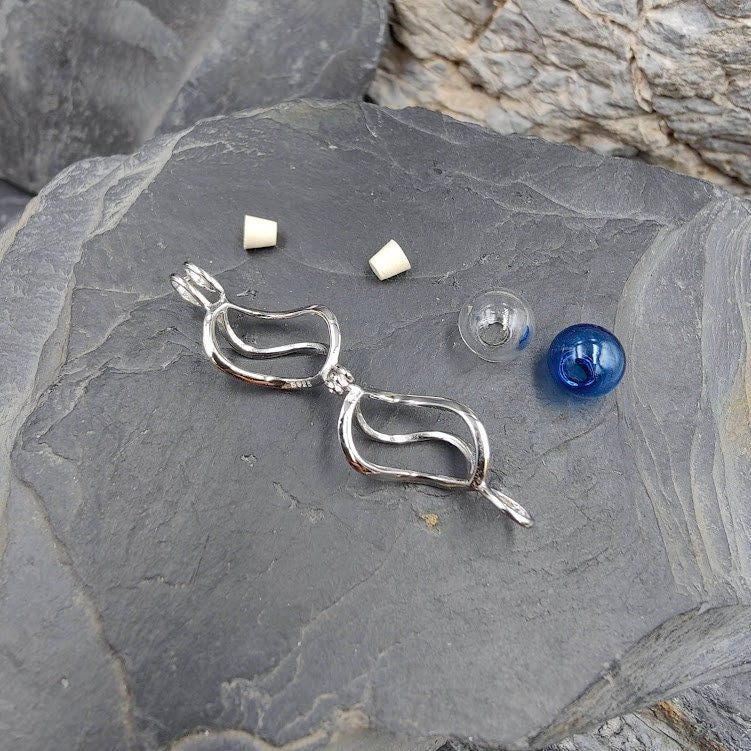 Einzelteile des Engelrufer DIY-Sets auf Schieferstein: silberner Käfig-Anhänger, blaue Glasperle, Glasphiole und Korken – Gedenkschmuck von Zinou Minimanufaktur