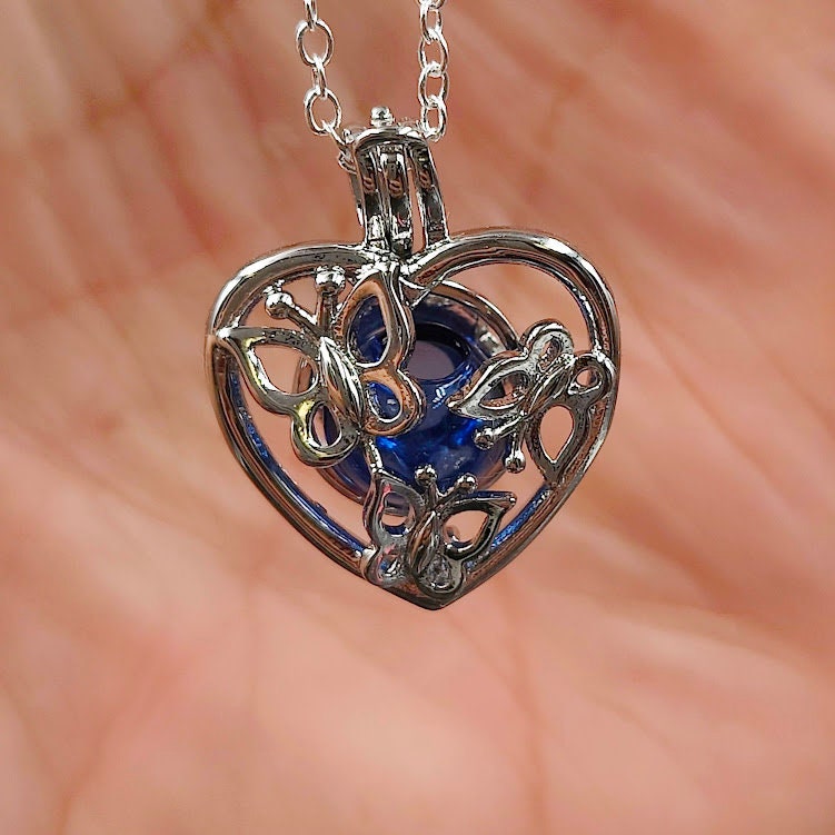 Silberner Herz-Käfig-Anhänger mit Schmetterlingsmotiv und blauer Glasperle in der Handfläche gehalten, Nahaufnahme – Gedenkschmuck zum selber Füllen von Zinou Minimanufaktur
