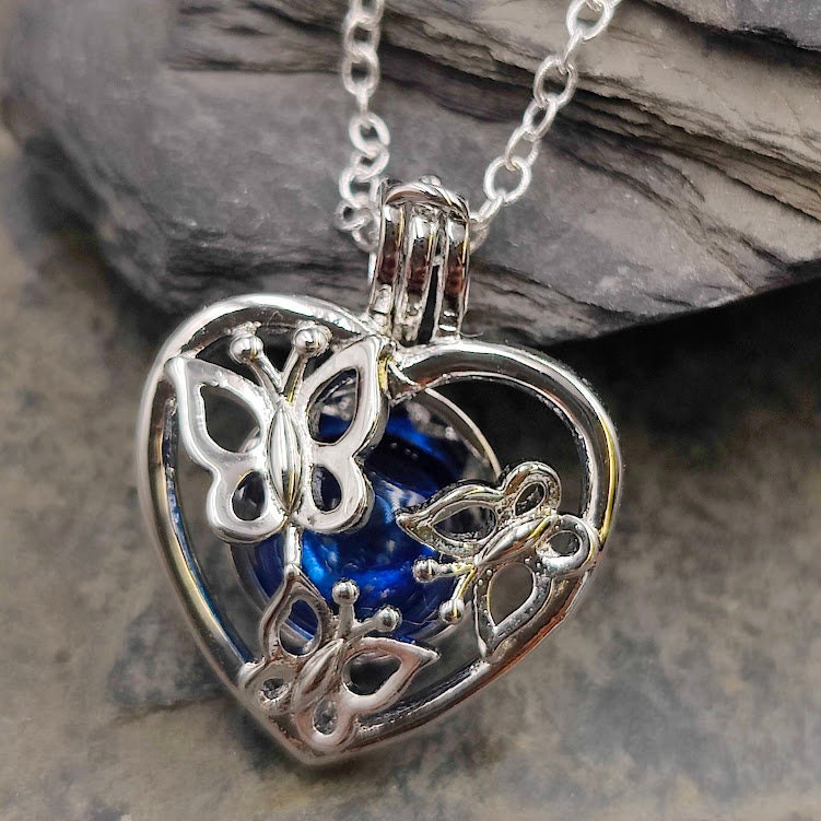 Silberner Herz-Käfig-Anhänger mit Schmetterlingsmotiv und blauer Glasperle auf Schieferstein vor Felsen, Nahaufnahme – Gedenkschmuck zum selber Füllen von Zinou Minimanufaktu