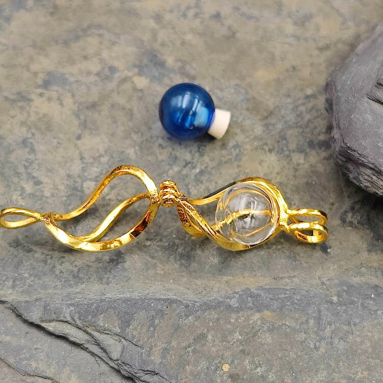 Goldener Spiralkäfig-Anhänger mit Glasperle zum Befüllen auf Schieferstein, blaue Perle daneben – Gedenkschmuck von Zinou Minimanufaktur