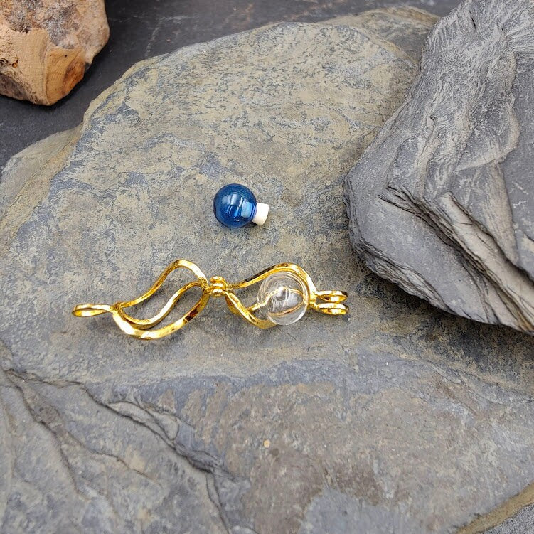 Goldener Spiralkäfig-Anhänger mit Glasperle zum Befüllen auf Schieferstein, blaue Perle daneben – Gedenkschmuck von Zinou Minimanufaktur