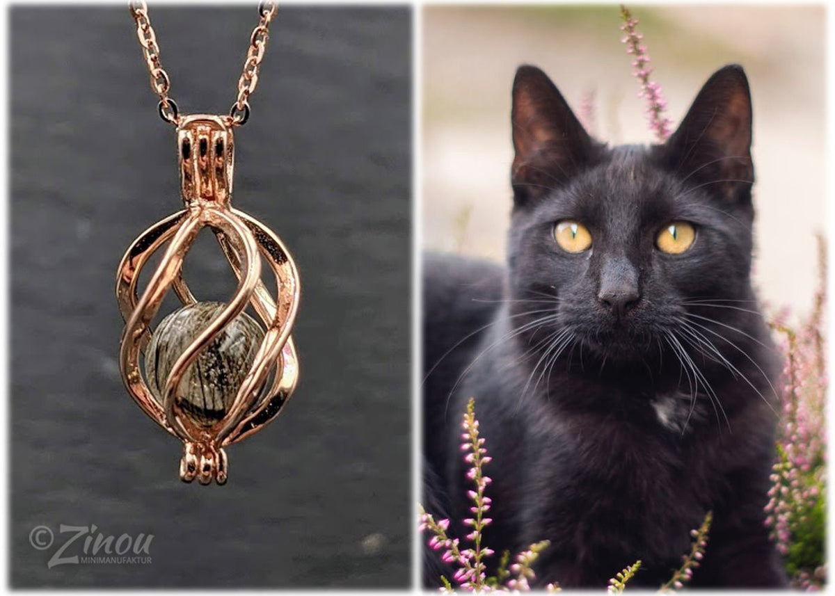 Rosegoldener Engelrufer-Käfig mit dunkler Fellperle an Rosegoldkette auf Schieferstein, schwarze Katze im Hintergrund – Tierhaarschmuck von Zinou Minimanufaktur