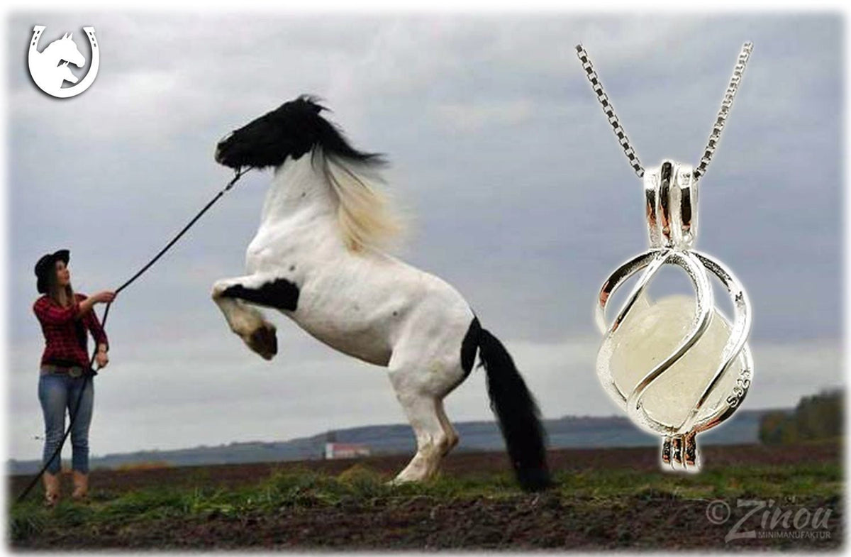 Silberner Engelrufer-Käfig mit weißer Fellperle an Silberkette, steigendes Pferd mit Reiterin im Hintergrund – Tierhaarschmuck von Zinou Minimanufaktur
