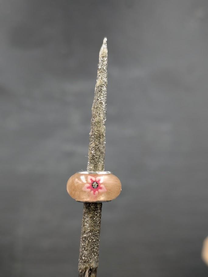 Handgefertigte Harzperle gefüllt mit Tierhaaren und verziert mit einer rosa Blüte, aufgefädelt auf einen rustikalen Holzstab vor grauem Hintergrund