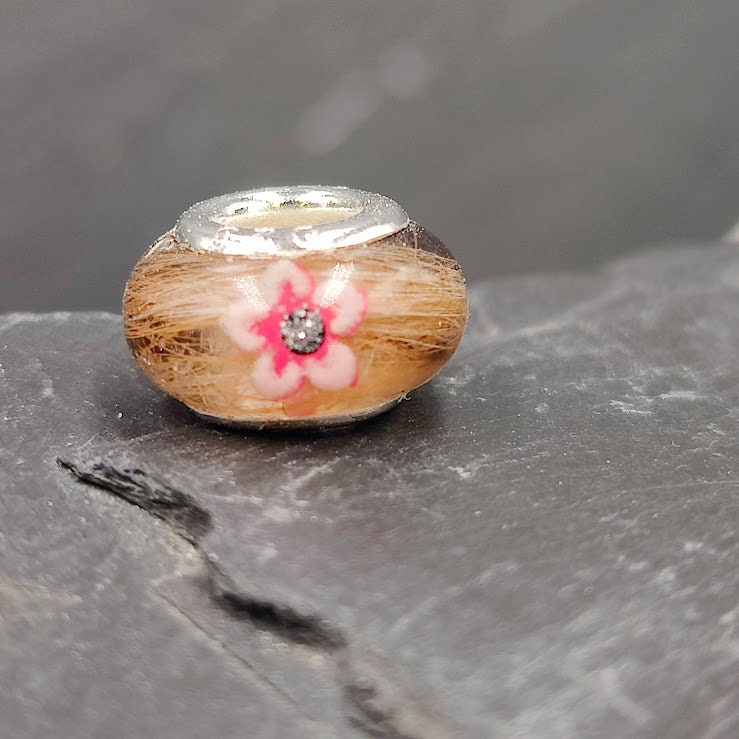 Handgefertigte Harzperle gefüllt mit braunem Tierhaarfell, verziert mit einer rosa Blüte und Glitzerstein, passende Öse für Bettelarmbänder