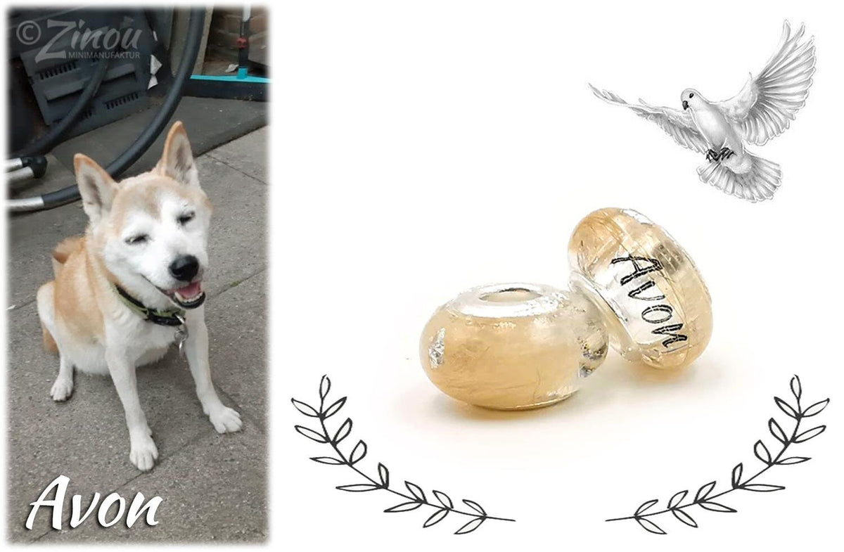 Zwei helle Gedenkperlen aus Harz mit dem Namen Avon, daneben ein Foto des weißen Hundes Avon sowie Zeichnungen einer Friedenstaube und eines Lorbeerkranzes