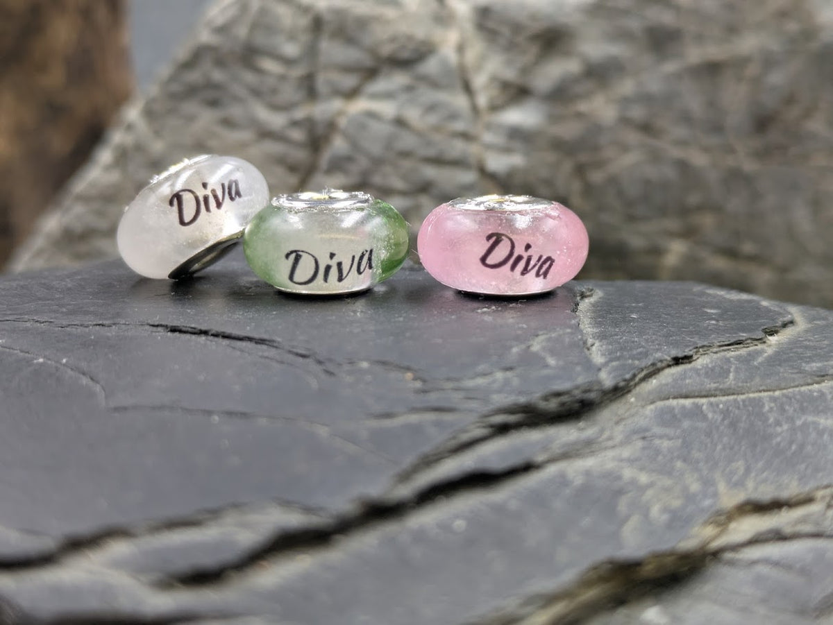 Drei Gedenkperlen gefüllt mit Asche in weiß, grün und rosa mit dem Namensaufdruck Diva, platziert auf dunklem Schiefergestein
