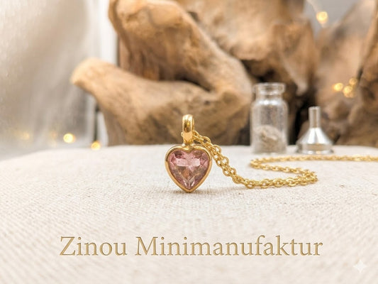 Goldene Halskette mit Herzanhänger und rosa Strassstein als befüllbare Mini-Urne, mit Befülltrichter und Glasfläschchen im Hintergrund auf hellem Untergrund