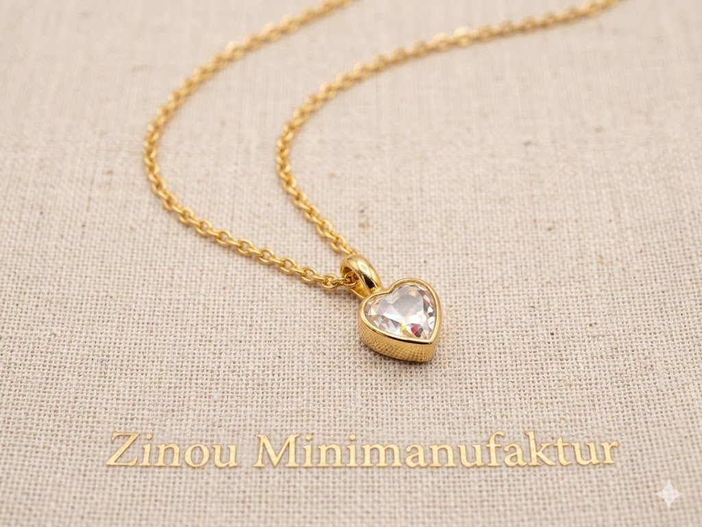 Nahaufnahme einer goldenen Halskette mit Herzanhänger und weißem Kristall als befüllbare Mini-Urne, liegend auf hellem Stoff mit Zinou Minimanufaktur Schriftzug
