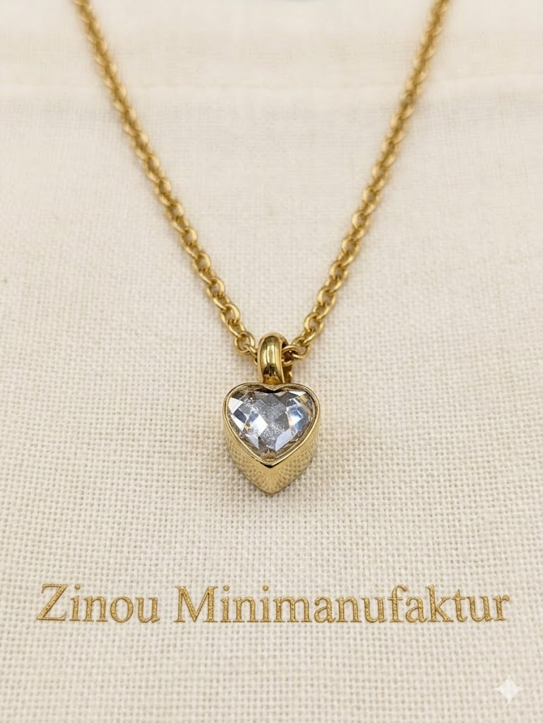 Goldene Halskette mit Herzanhänger und gefasstem weißem Kristall als befüllbare Mini-Urne, präsentiert auf hellem Stoff mit Zinou Minimanufaktur SchriftzugHerzurne Anhänger aus Edelstahl Gold Urnenschmuck für Asche 
