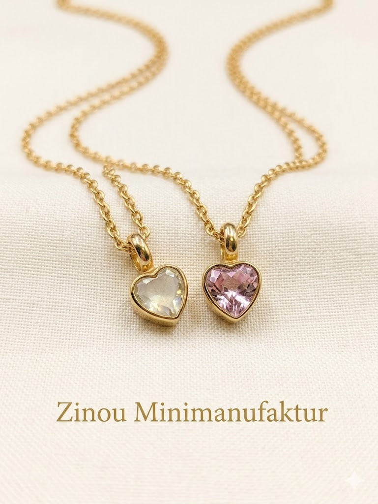 Zwei goldene Halsketten mit befüllbaren Herzanhängern, eine mit weißem und eine mit rosa Kristall, nebeneinander liegend auf hellem Stoff mit Zinou Minimanufaktur Schriftzug