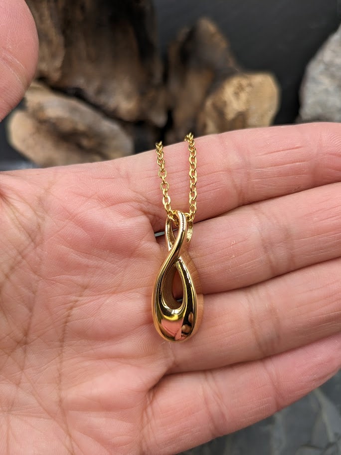 Goldener Infinity-Urnenanhänger an Goldkette hängend vor grauem Hintergrund mit Schieferstein, Frontansicht – Gedenkschmuck von Zinou Minimanufaktur