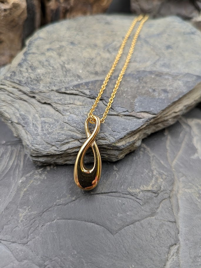 Goldener Infinity-Urnenanhänger an Goldkette hängend vor grauem Hintergrund mit Schieferstein, Frontansicht – Gedenkschmuck von Zinou Minimanufaktur