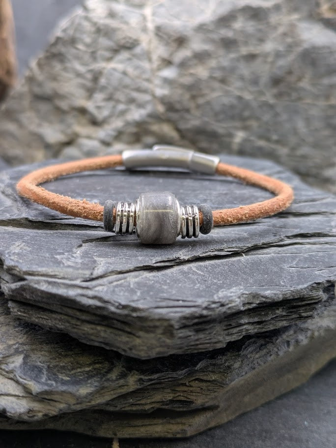 Hellbraunes Lederarmband mit eckiger Harzperle gefüllt mit grauem Katzenfell, liegend auf einem dunklen Schieferstein