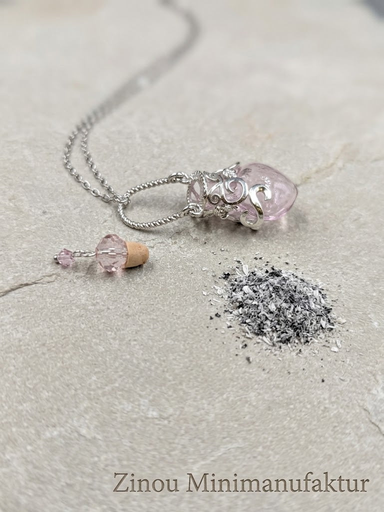 Geöffnete rosa Herz-Glasphiole mit abgezogenem Korken neben einer kleinen Portion Asche – zeigt die Befüllmöglichkeit für Gedenkschmuck, Zinou Minimanufaktur