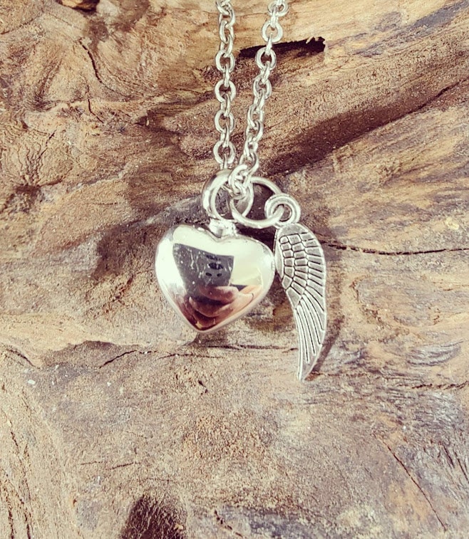 Silberne Herz-Urne mit Flügel-Charm an Silberkette auf Treibholz, Draufsicht – Gedenkschmuck von Zinou Minimanufaktur
