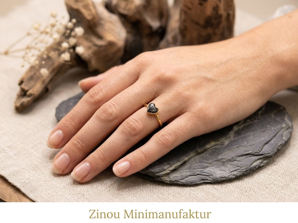 Hand auf Schieferstein trägt goldenen Herzring mit schwarzem Asche-Einschluss vor Treibholz – Gedenkring von Zinou Minimanufaktur