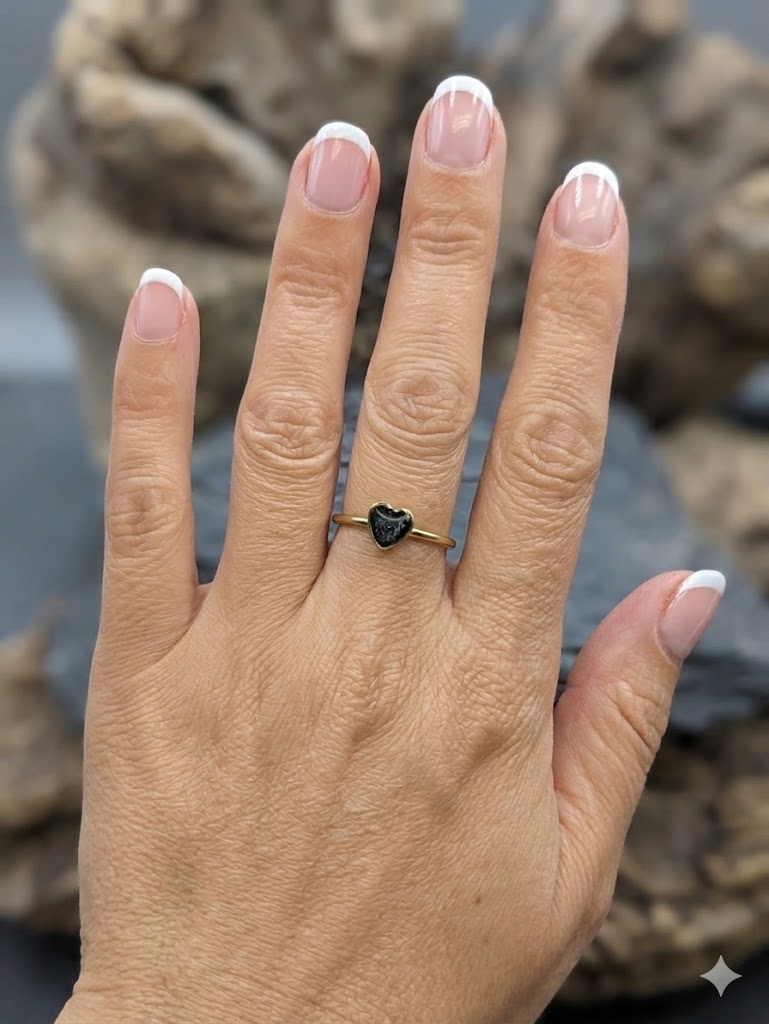 Hand trägt goldenen Herzring mit schwarzem Asche-Einschluss vor Treibholz auf Schieferstein – Gedenkring von Zinou Minimanufaktur