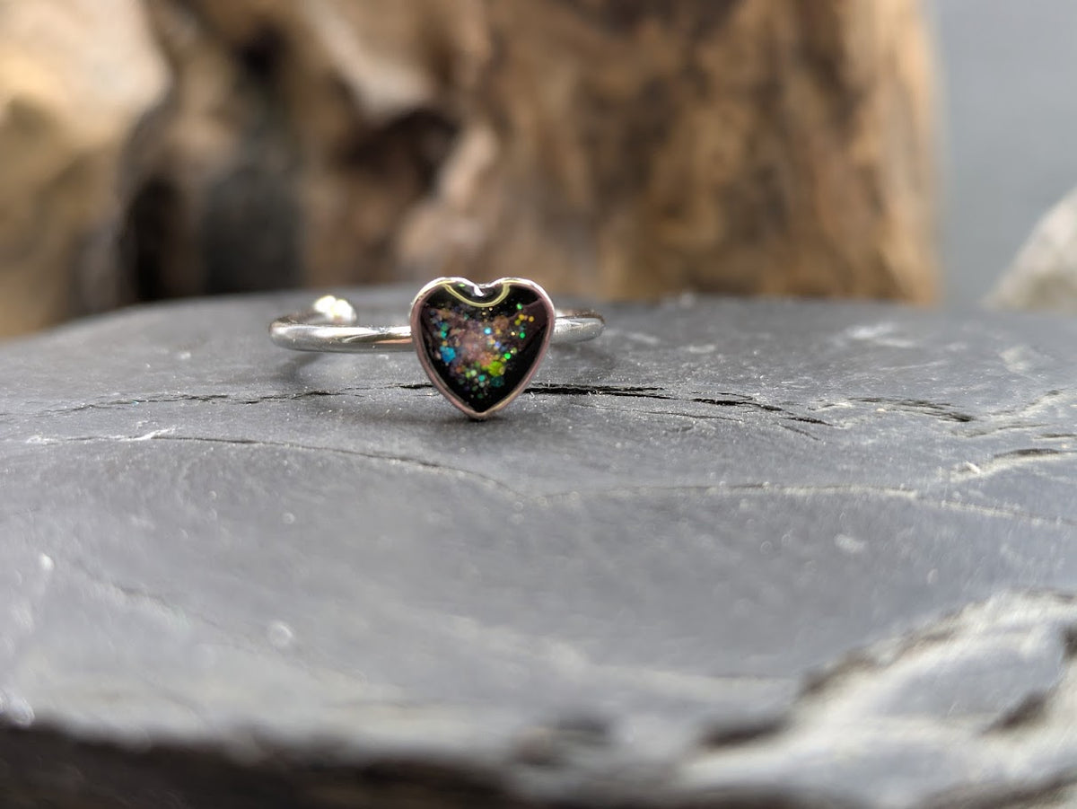 Silberner Herzring mit schwarzem Asche- und buntem Opal-Glitter-Einschluss auf Schieferstein vor Treibholz, Nahaufnahme – Gedenkring von Zinou Minimanufaktur