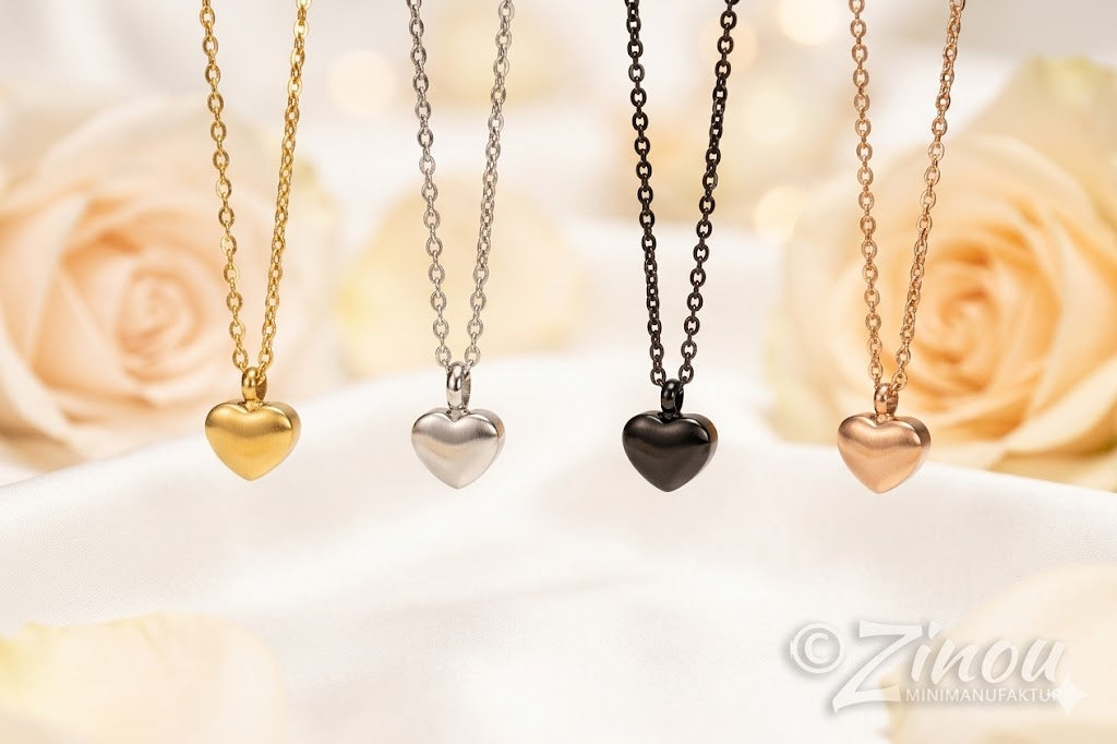 Vier befüllbare Herzurnenketten in den Farben Gold, Silber, Schwarz und Roségold nebeneinander aufgereiht vor hellen Rosenblüten mit Zinou Minimanufaktur Wasserzeichen