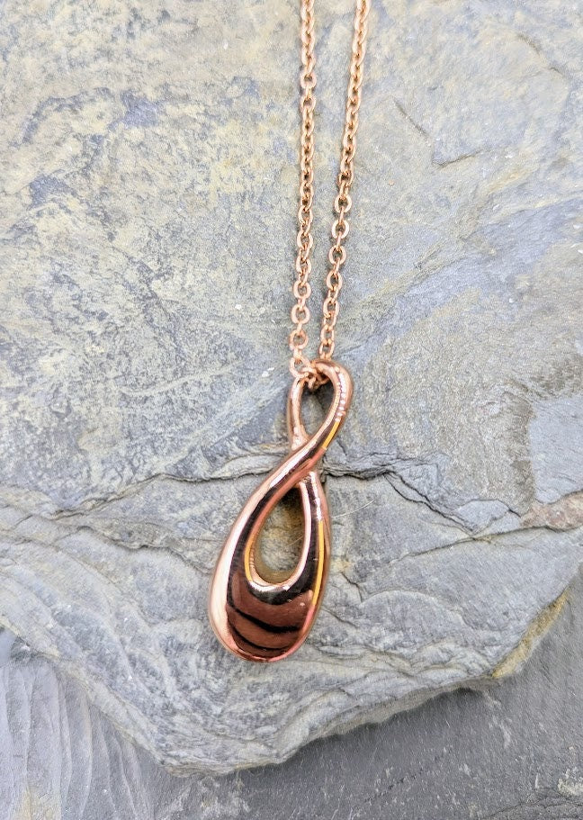 Rosefarbiger  Infinity-Urnenanhänger an Goldkette hängend vor grauem Hintergrund mit Schieferstein, Frontansicht – Gedenkschmuck von Zinou Minimanufaktur