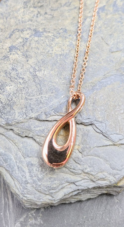 Goldener Infinity-Urnenanhänger an Goldkette hängend vor grauem Hintergrund mit Schieferstein, Frontansicht – Gedenkschmuck von Zinou Minimanufaktur