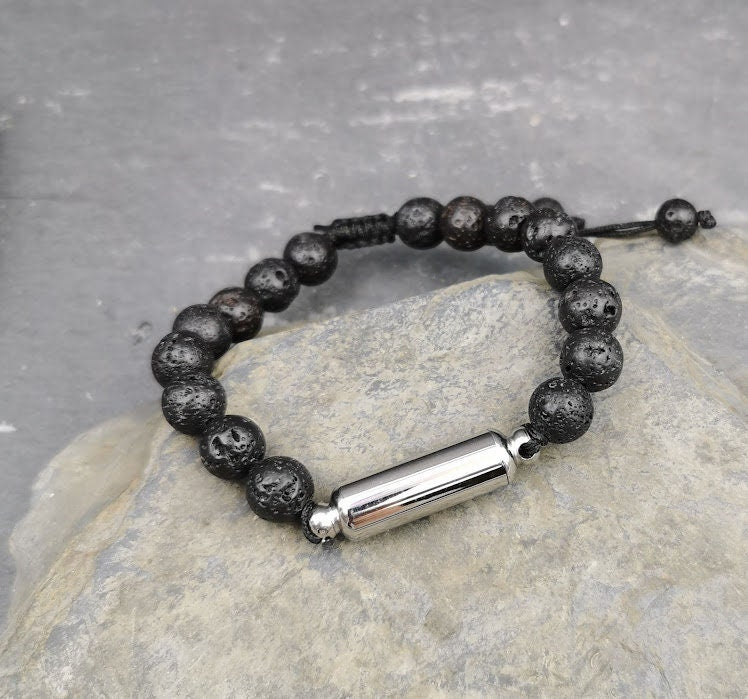Männerarmband aus schwarzen Lavaperlen mit silberner Miniurne auf hellem Marmor, erhöhte Perspektive – Gedenkschmuck von Zinou Minimanufaktur