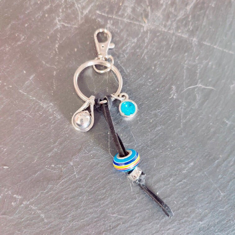 Schlüsselanhänger mit silberner Tränen-Miniurne, türkisem Strassstein-Charm und bunter Ringperle auf schwarzer Lederkordel auf Schieferstein – Gedenkschmuck von Zinou Minimanufaktur