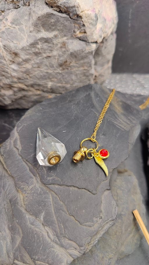 Pendel-Urne aus Bergkristall in Spitzenform mit Flügel-Charm und rotem Kristallstein an Goldkette auf Schieferstein zwischen Felsen – Gedenkschmuck von Zinou Minimanufaktur