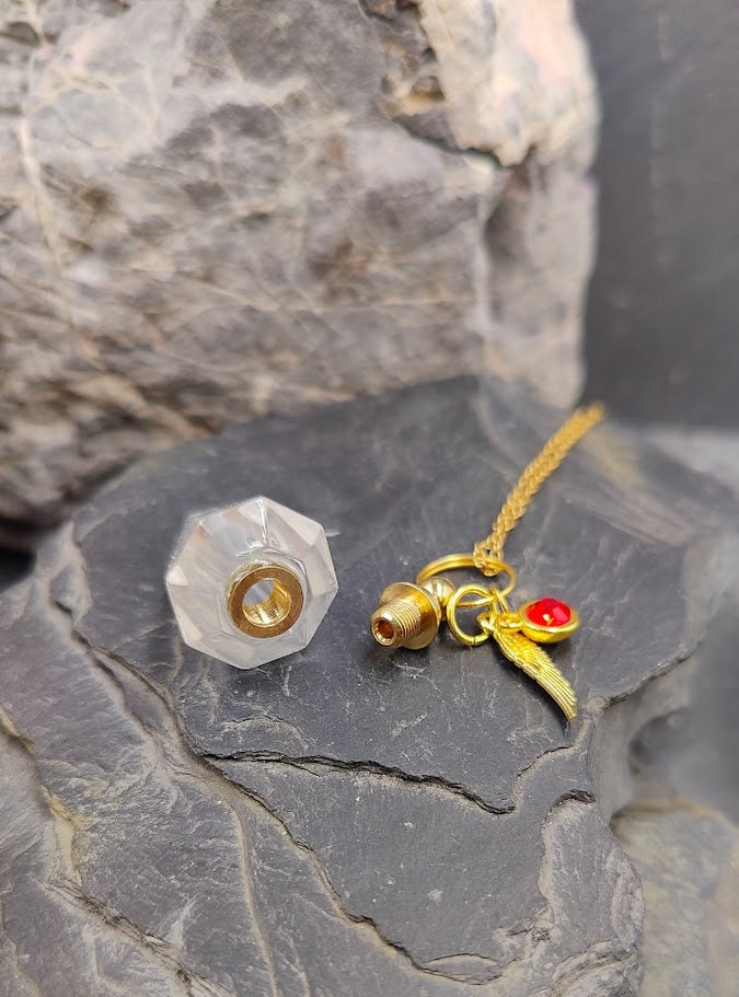 Pendel-Urne aus Bergkristall in Spitzenform mit Flügel-Charm und rotem Kristallstein an Goldkette geöffnet auf Schieferstein zwischen Felsen – Gedenkschmuck von Zinou Minimanufaktur