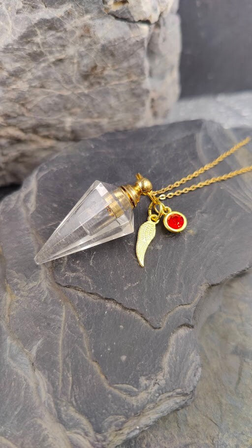 Pendel-Urne aus Bergkristall in Spitzenform mit Flügel-Charm und rotem Kristallstein an Goldkette auf Schieferstein, seitliche Ansicht – Gedenkschmuck von Zinou Minimanufaktur