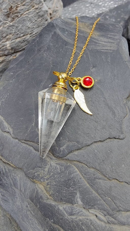 Pendel-Urne aus Bergkristall in Spitzenform mit goldener Fassung, Flügel-Charm und rotem Kristallstein an Goldkette auf Schieferstein – Gedenkschmuck von Zinou Minimanufaktur