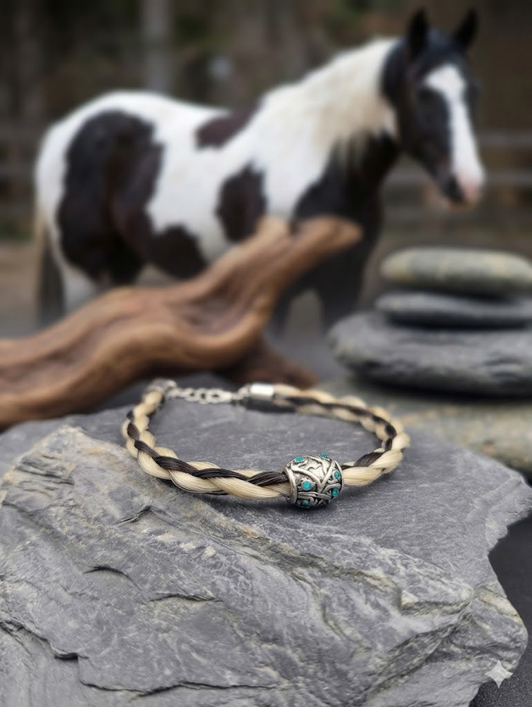Geflochtenes hell-dunkelbraunes Pferdehaararmband mit silberner Perle und türkisen Steinen auf Schieferstein, schwarz-weißes Pferd im Hintergrund – Pferdehaarschmuck von Zinou Minimanufaktur