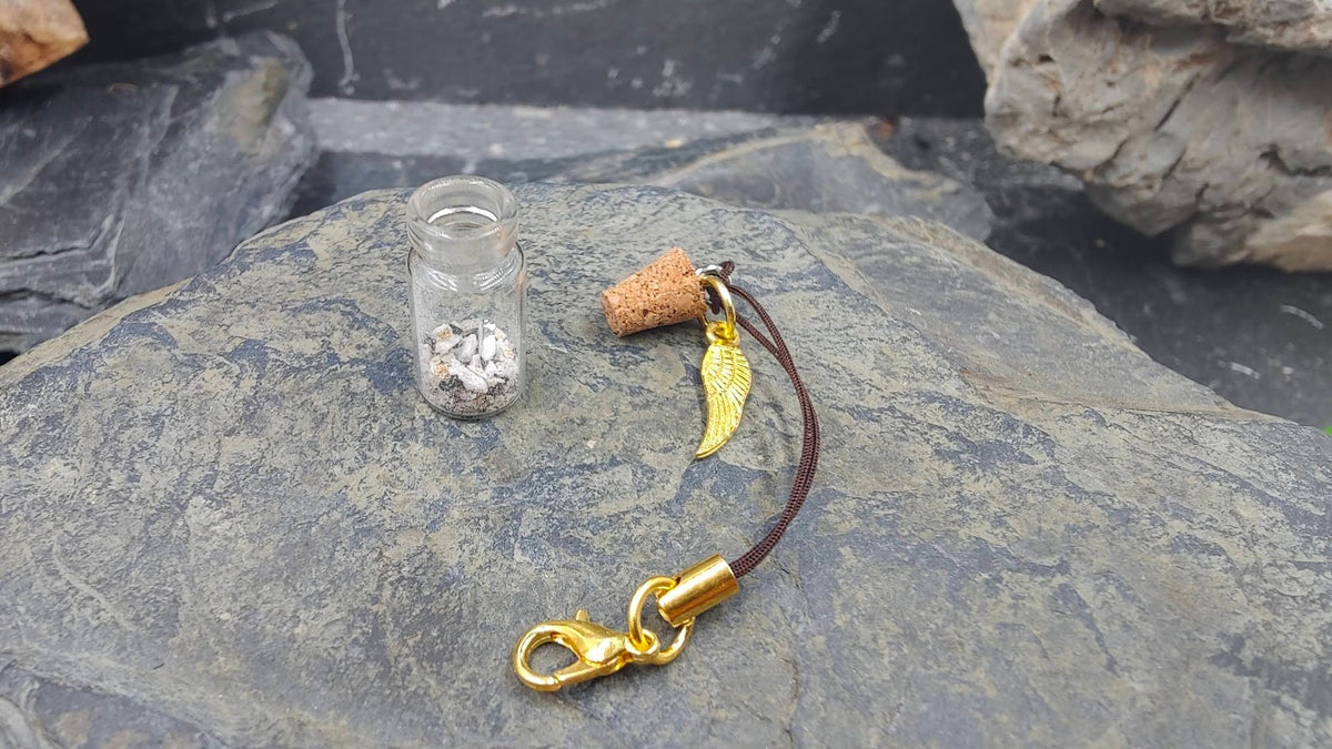 Einzelteile der Phiole Gold mit Flügelcharm auf Schieferstein: geöffnetes Glasfläschchen mit Fellinhalt, Korkenstopfen, goldener Flügel-Charm und brauner Kordel mit Karabiner – DIY Gedenkschmuck von Zinou Minimanufaktur