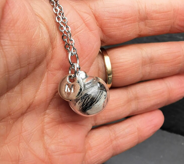 Glasphiole mit Tierhaar und silbernem Buchstaben-Charm M an Silberkette in der Handfläche gehalten, Nahaufnahme – personalisierter Gedenkschmuck von Zinou Minimanufaktur