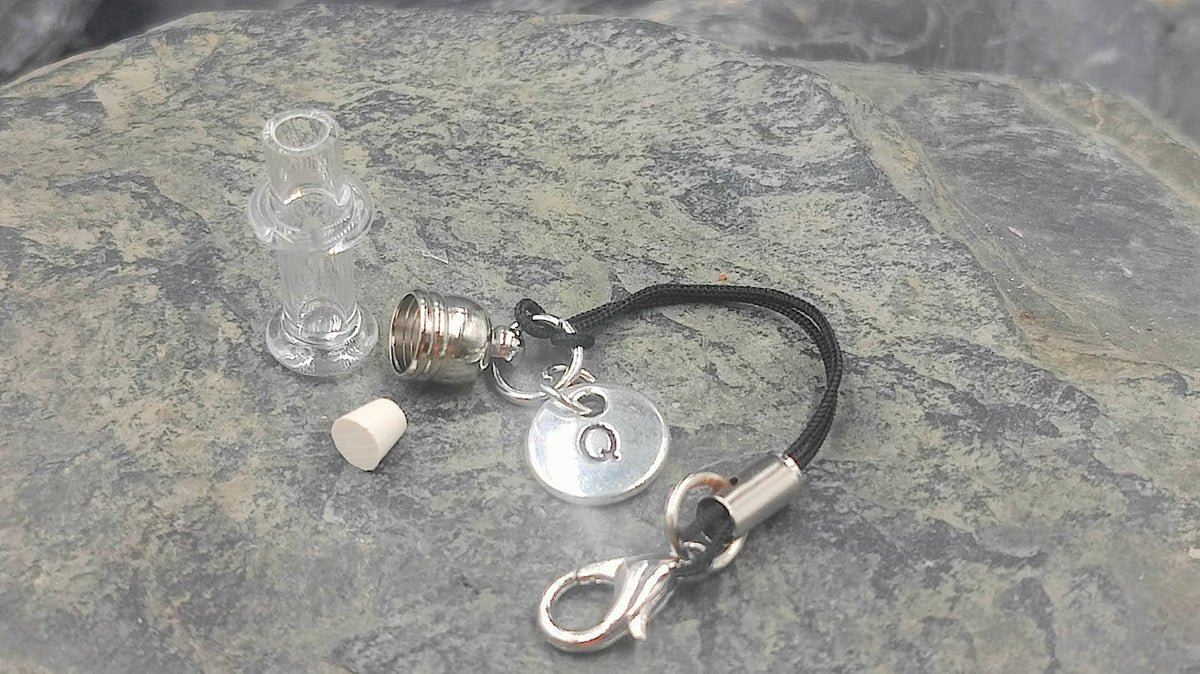 Einzelteile des Phiole Mini Fläschen Sets auf Schieferstein: leeres Glasfläschchen, Korken, silberne Kappe, Buchstaben-Charm Q und schwarzer Kordel mit Karabiner – Gedenkschmuck von Zinou Minimanufaktur