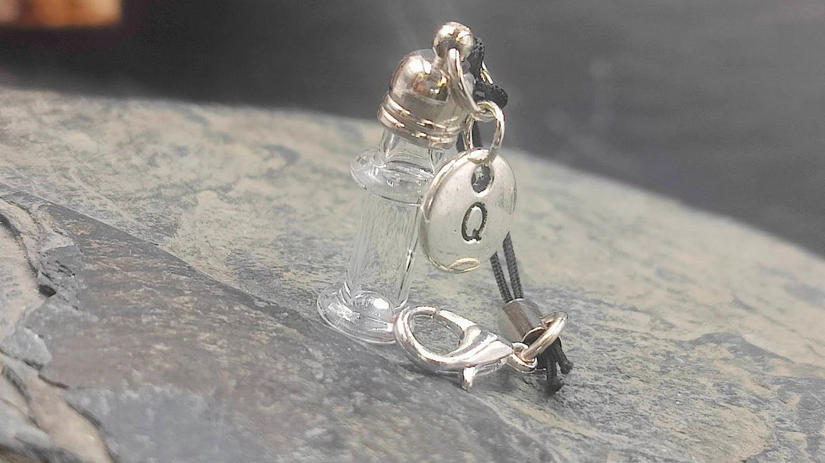 Leeres Mini Glasfläschchen mit silbernem Buchstaben-Charm Q an schwarzem Kordel-Schlüsselanhänger auf Schieferstein neben Treibholz – Gedenkschmuck von Zinou Minimanufaktur