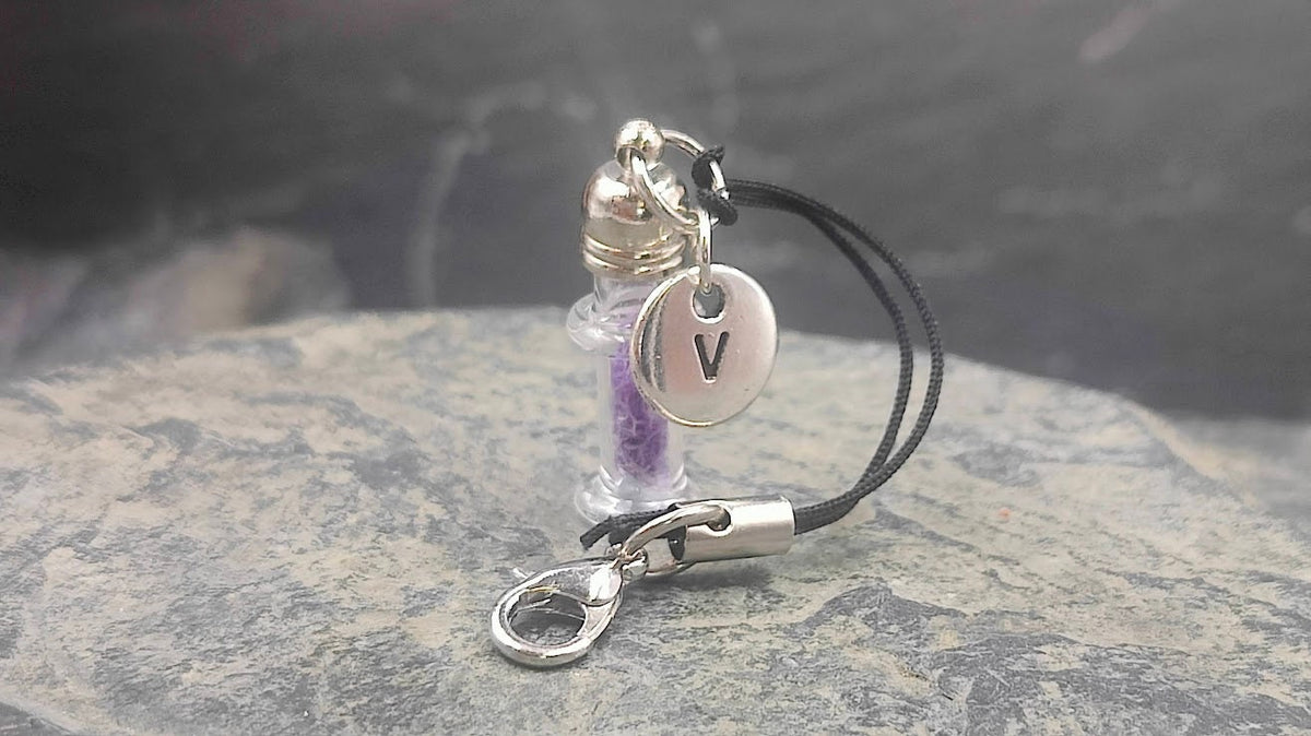 Mini Glasfläschchen mit lilafarbenem Inhalt und silbernem Buchstaben-Charm V an schwarzem Kordel-Schlüsselanhänger auf Schieferstein – Gedenkschmuck von Zinou Minimanufaktur