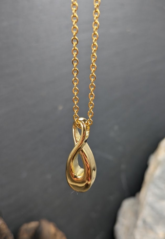 Goldener Infinity-Urnenanhänger an Goldkette hängend vor grauem Hintergrund mit Schieferstein, Frontansicht – Gedenkschmuck von Zinou Minimanufaktur