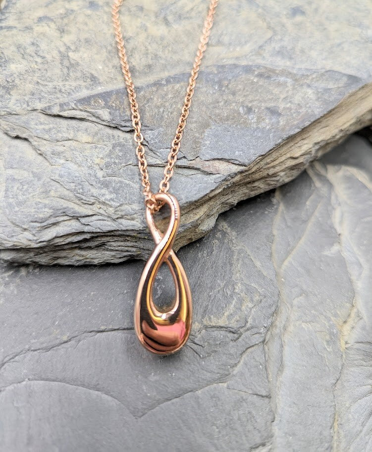 Roségoldener Infinity-Urnenanhänger an Roségoldkette hängend auf Schieferstein, seitliche Ansicht – Gedenkschmuck von Zinou Minimanufaktur